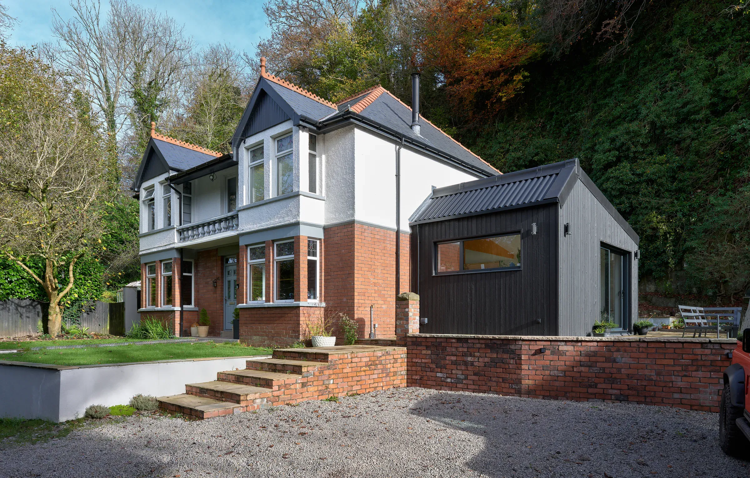 Front of the house with its new black-clad extension, gravel drive and the wooded valley behind